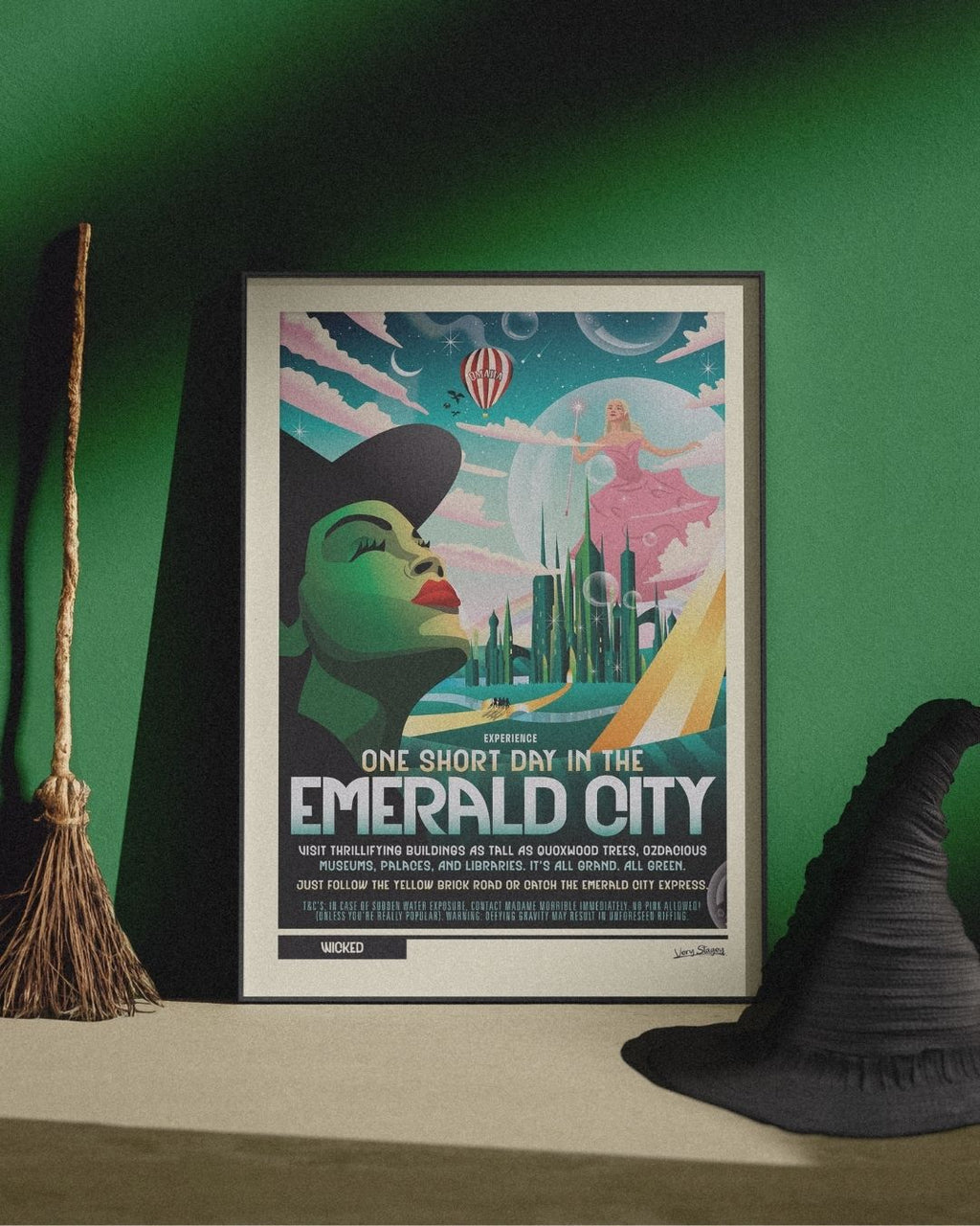 One Short Day Wicked poster in a black frame against a green wall, with a broom and a witch's hat