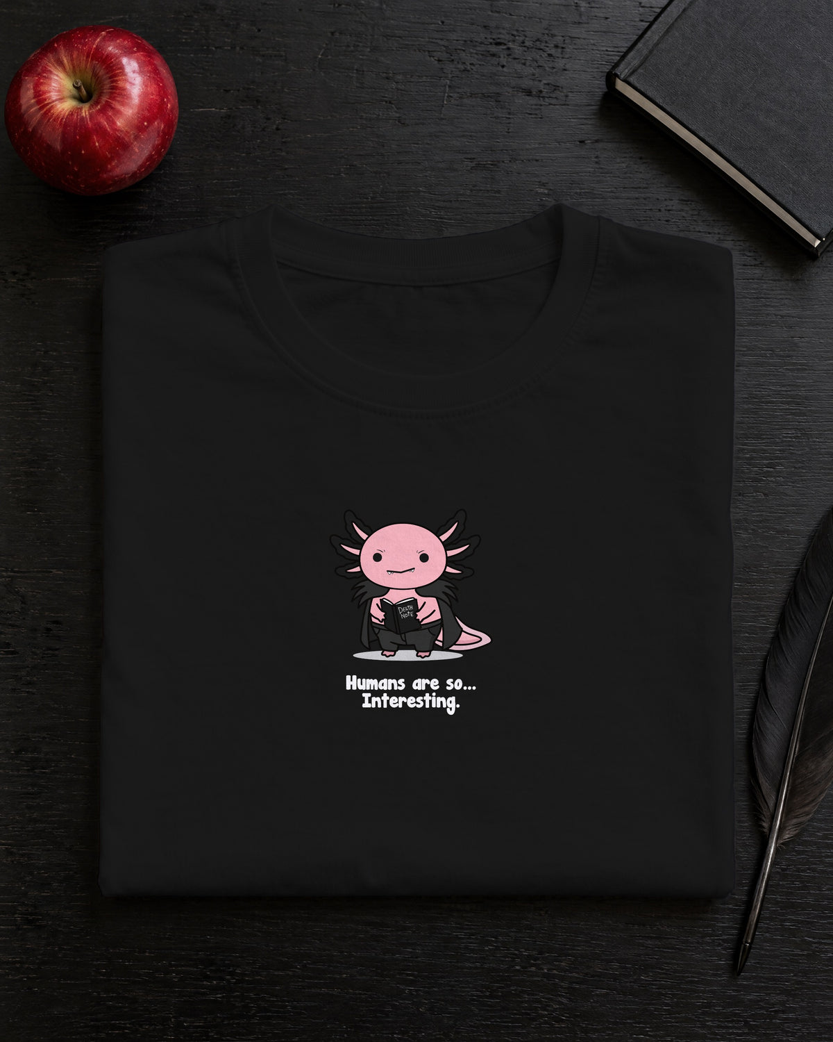 Humans Are So Interesting t-shirt in Black, folded on a black desk with an apple, a notebook and a quill