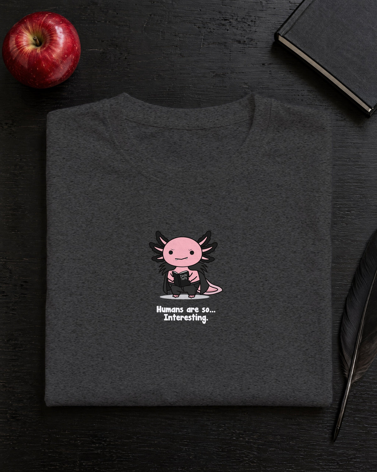 Humans Are So Interesting t-shirt in Dark Heather Grey, folded on a black desk with an apple, a notebook and a quill