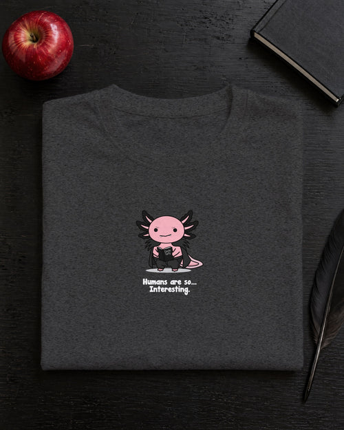 Humans Are So Interesting t-shirt in Dark Heather Grey, folded on a black desk with an apple, a notebook and a quill
