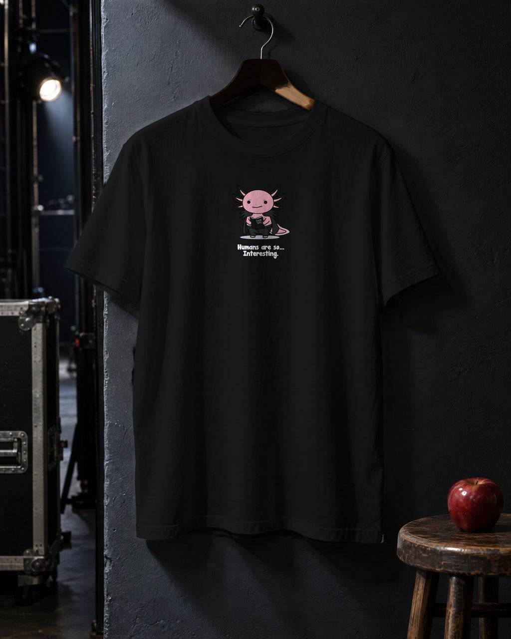 Humans Are So Interesting t-shirt in Black on a hanger against a dark backstage wall, with a red apple