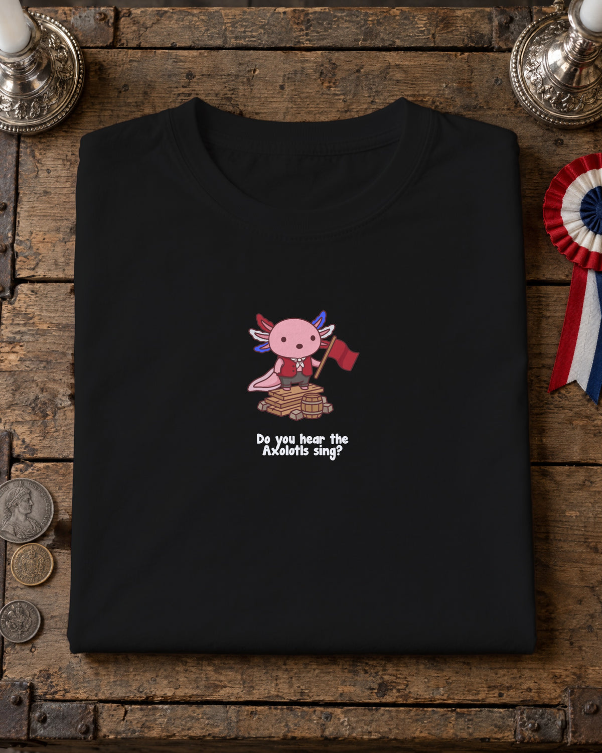 Do You Hear the Axolotls Sing t-shirt in Black, folded on a crate lid with silver candlesticks and a tricolour rosette