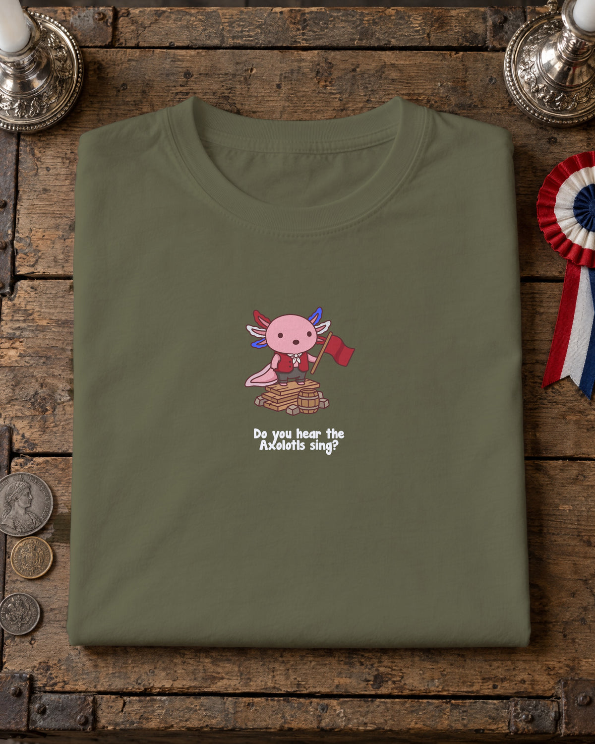 Do You Hear the Axolotls Sing t-shirt in Khaki, folded on a crate lid with silver candlesticks and a tricolour rosette